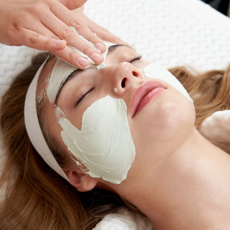 Cosmetologist applying rejuvenating clay facial mask on a female face in beauty salon. Facial treatment concept
